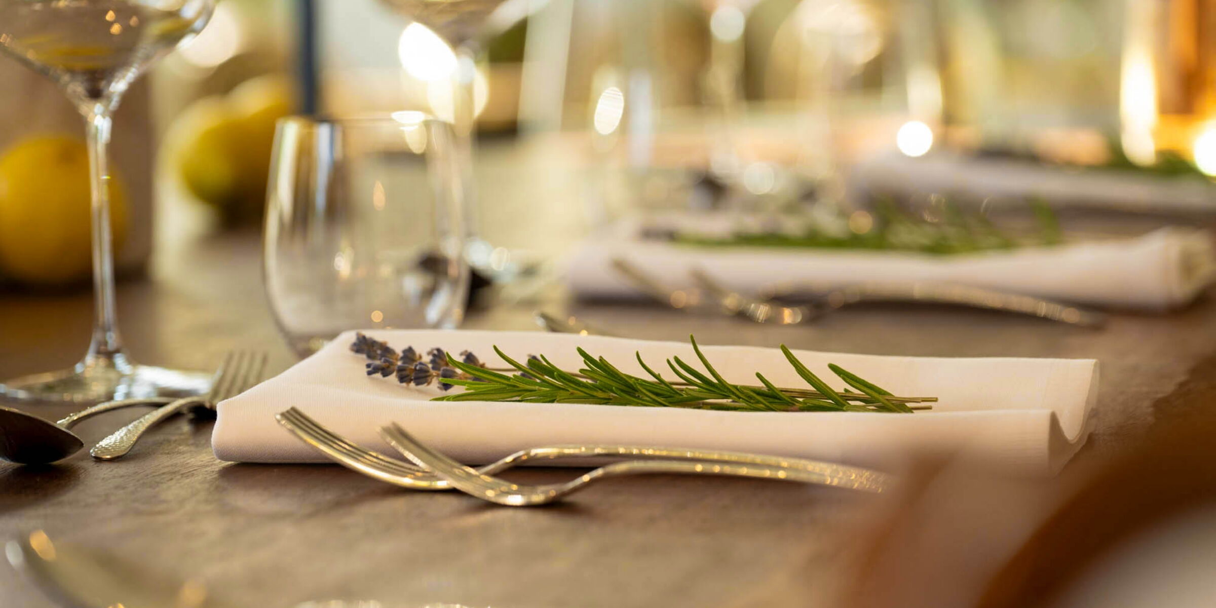 Ein elegant gedeckter Tisch mit gefalteten Stoffservietten, auf denen Lavendel und Rosmarin liegen. Im Vordergrund sind Gabeln und im Hintergrund Gläser und verschwommene Lichter sichtbar.