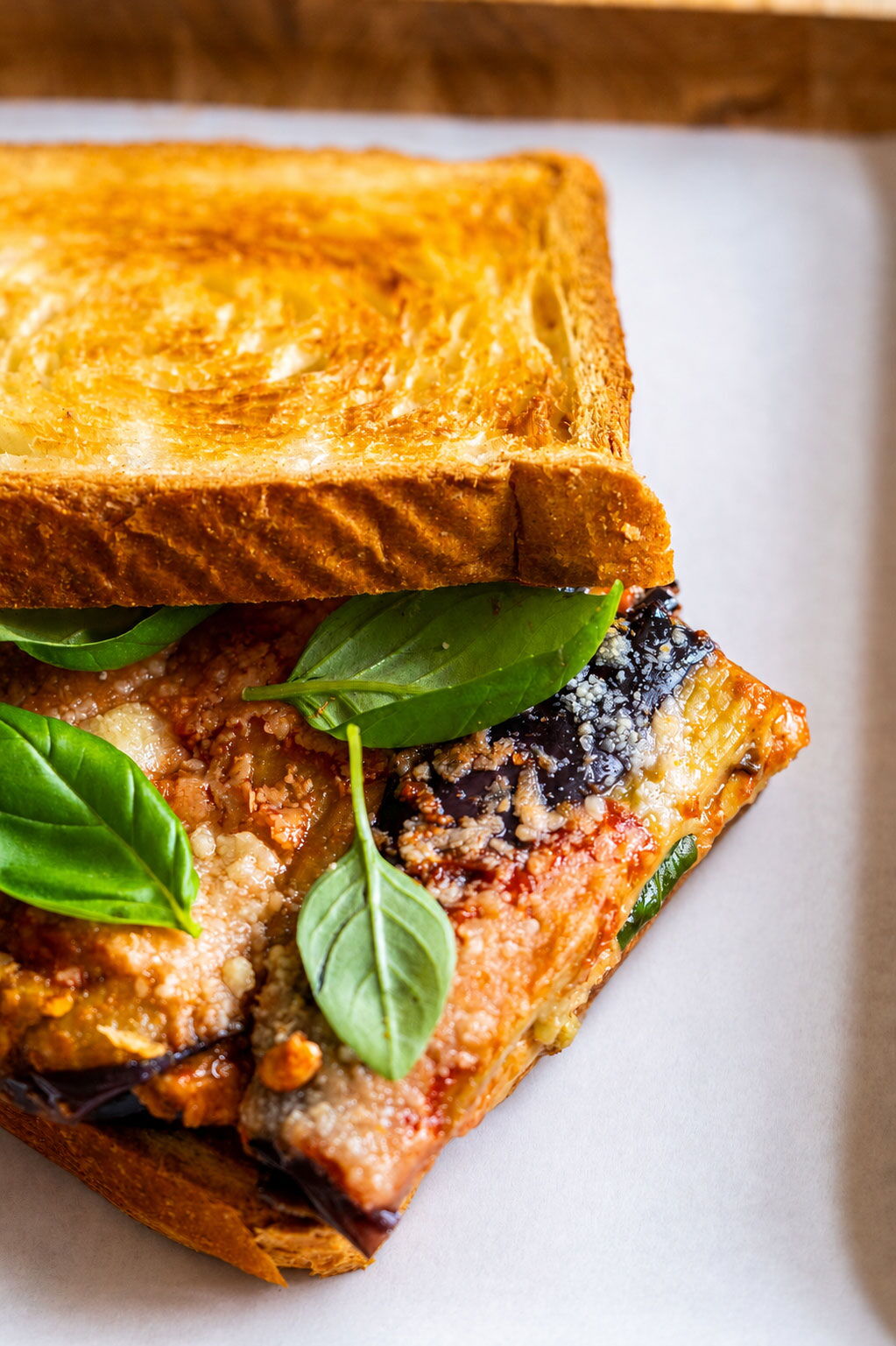 Ein Sandwich mit goldbraun getoastetem Brot, gefüllt mit einer Schicht Melanzani, Tomatensauce, Parmesan und frischen Basilikumblättern auf einem weißen Teller.