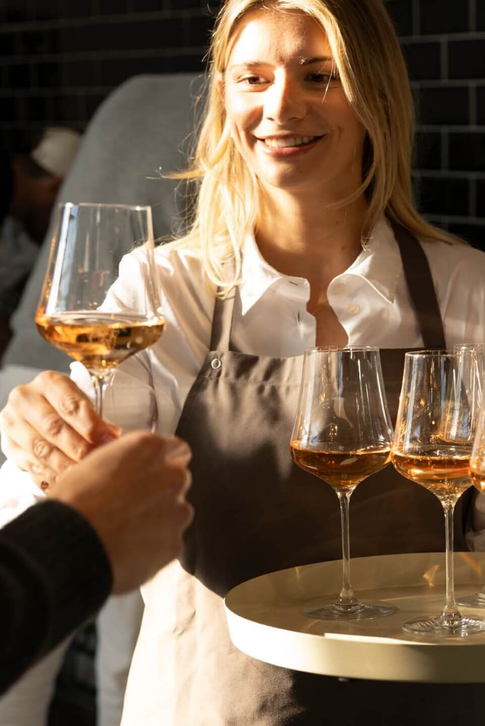 Eine Frau in einer Schürze reicht einem Gast ein Glas Roséwein. Sie hält ein Tablett mit mehreren Gläsern. Die Szene ist in warmem Licht gehalten. Im Hintergrund ist ein unscharfer dunkler Bereich.