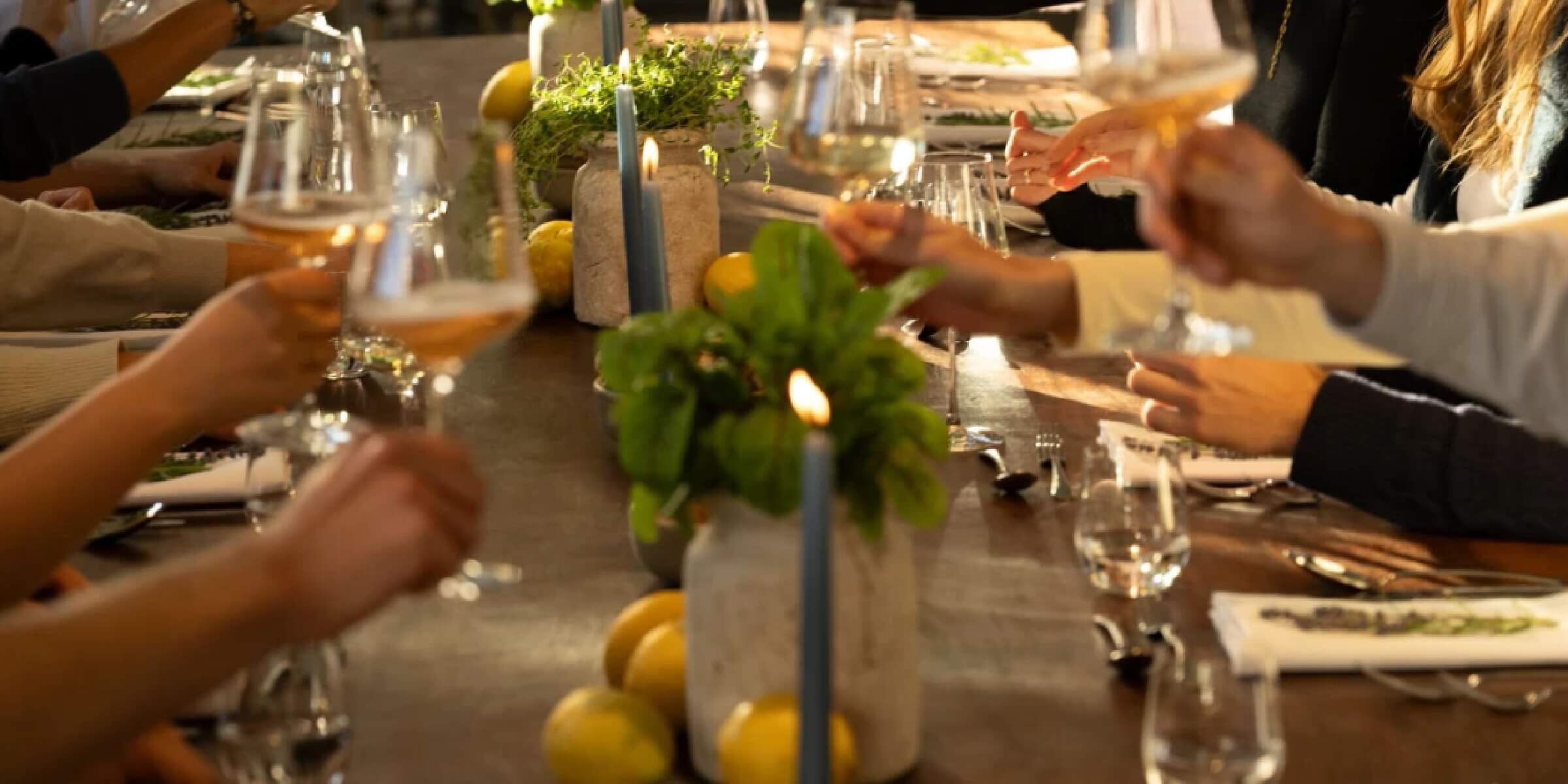 Eine Gruppe von Menschen sitzt an einem festlichen Tisch und erhebt Gläser, um anzustoßen. Der Tisch ist mit Kerzen, grünen Pflanzen und Zitronen dekoriert. Die Lichtstimmung ist warm und einladend.